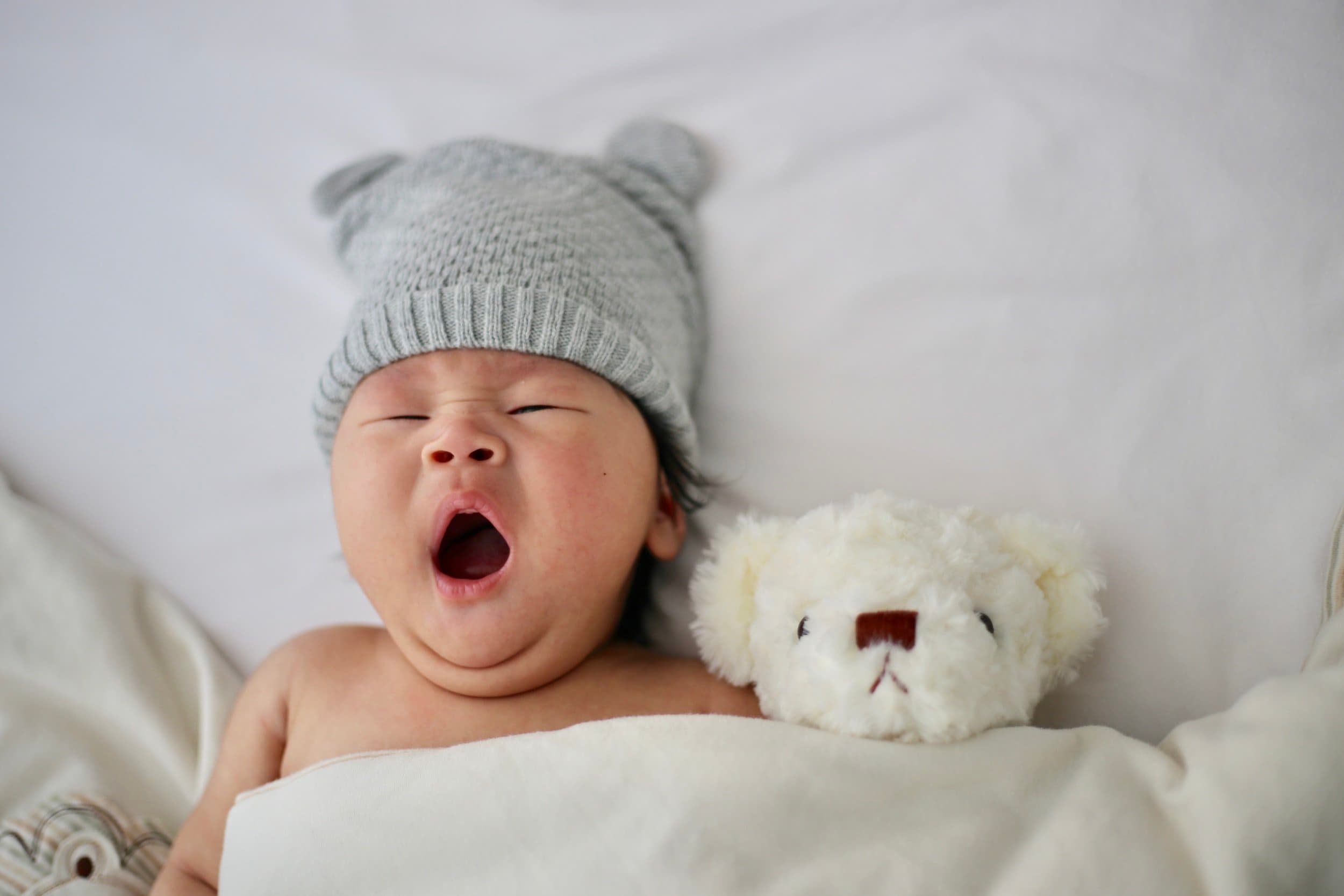 A baby wearing a hat and yawning beside a teddy in a bed.